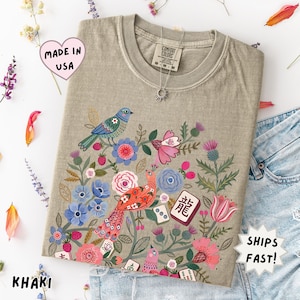 May include: A khaki t-shirt featuring a graphic print of colorful birds, various flowers, and mahjong tiles. The shirt has a crew neckline and a Comfort Colors brand label at the inner collar. The design includes blue, pink, and red floral elements.