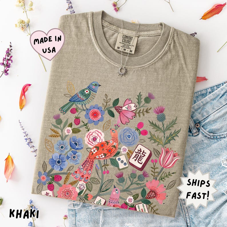 May include: A khaki t-shirt featuring a graphic print of colorful birds, various flowers, and mahjong tiles. The shirt has a crew neckline and a Comfort Colors brand label at the inner collar. The design includes blue, pink, and red floral elements.