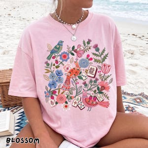 May include: A light pink t-shirt featuring a colorful graphic print of birds, various flowers, and mahjong tiles. The design includes a blue bird, an orange bird, a pink bird, and several mahjong tiles with Chinese characters and dots.