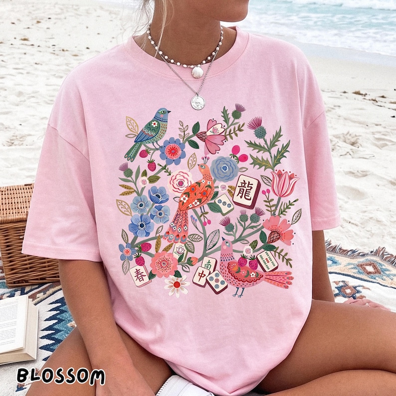 May include: A light pink t-shirt featuring a colorful graphic print of birds, various flowers, and mahjong tiles. The design includes a blue bird, an orange bird, a pink bird, and several mahjong tiles with Chinese characters and dots.