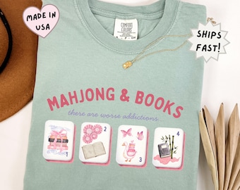 May include: A sage green t-shirt featuring the pink text "MAHJONG & BOOKS" and "there are worse addictions..." above four white mahjong tiles. The tiles display illustrations of stacked books, pink rosettes, a goose with books, and a book with bamboo.