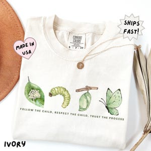 May include: An ivory t-shirt featuring a watercolor illustration of the butterfly life cycle, including eggs on a leaf, a caterpillar, a chrysalis, and a butterfly. The text reads, "FOLLOW THE CHILD, RESPECT THE CHILD, TRUST THE PROCESS."