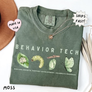 May include: A moss green t-shirt featuring the text BEHAVIOR TECH in light green letters. Below the text are watercolor illustrations of a leaf with eggs, a caterpillar, a chrysalis, and a butterfly. The text NURTURE GROWTH, SUPPORT DEVELOPMENT, CELEBRATE PROGRESS is printed at the bottom.