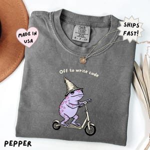 May include: A charcoal gray crewneck t-shirt featuring a graphic of a light purple frog with pink heart spots wearing a tan pointed hat and riding a silver scooter. The text above the frog reads Off to write code in white lettering.