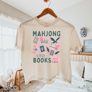 May include: Ivory-colored t-shirt with the words "MAHJONG AND BOOKS" in green. The shirt features a graphic design with a crown, dragon, and other elements in pink, blue, and black. The shirt is hanging on a wooden hanger.