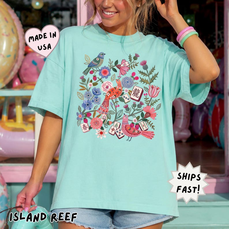 May include: A light turquoise t-shirt featuring a colorful graphic print of birds, flowers, and mahjong tiles. The design includes red, blue, and pink floral elements, two birds, and several mahjong tiles with Chinese characters. The shirt has short sleeves.