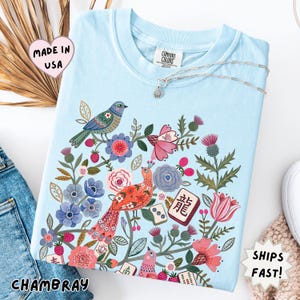 May include: A light blue t-shirt featuring a colorful graphic print of birds, various flowers, and mahjong tiles. The shirt has a crew neckline and a Comfort Colors brand label at the inner collar. The graphic includes red, pink, and blue floral elements.
