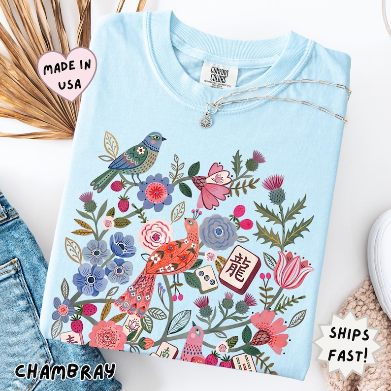 May include: A light blue t-shirt featuring a colorful graphic print of birds, various flowers, and mahjong tiles. The shirt has a crew neckline and a Comfort Colors brand label at the inner collar. The graphic includes red, pink, and blue floral elements.