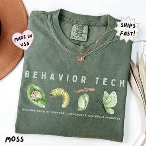 May include: Moss green t-shirt with the words "BEHAVIOR TECH" and a graphic of a caterpillar life cycle. The text "NURTURE GROWTH, SUPPORT DEVELOPMENT, CELEBRATE PROGRESS" is printed below the graphic. A silver necklace with a pendant is on the shirt.