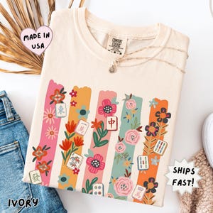May include: An ivory t-shirt featuring a graphic print of vertical pastel-colored stripes interspersed with various floral illustrations and mahjong tiles. The shirt has a crew neckline and a Comfort Colors brand label at the inner collar.
