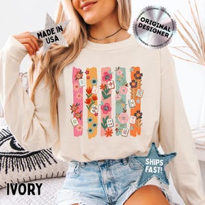 May include: An ivory long-sleeve sweatshirt featuring a graphic print of five vertical stripes in pink, orange, and teal. The design includes colorful floral illustrations and various Mahjong tiles with Chinese characters and symbols scattered throughout.