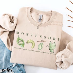 May include: An ivory-coloured sweatshirt featuring the word "MONTESSORI" in green, with illustrations of a leaf, caterpillar, chrysalis, and butterfly. The text "FOLLOW THE CHILD, RESPECT THE CHILD, TRUST THE PROCESS" is printed below the illustrations. A silver necklace is also visible.