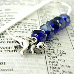 May include: A silver dinosaur charm bookmark with a string of faceted, deep blue beads. The bookmark is set against the pages of a book, with text visible in the background.