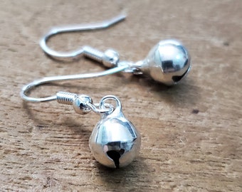 May include: A pair of silver dangle earrings with small bell charms. The bells have a rounded shape and a small opening at the top.