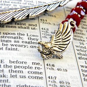 May include: A silver-colored metal bookmark with a feather design and an eagle charm, accented with burgundy beads. The bookmark rests on the open pages of a book, with visible text.