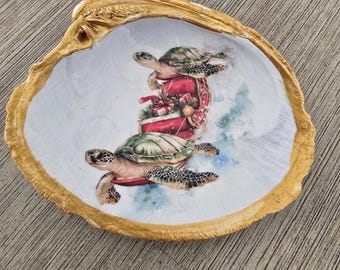 May include: A decorative seashell with a gold-painted rim and a painted interior featuring two sea turtles. The turtles are depicted with a red sack of gifts, creating a festive holiday theme. The shell is resting on a wooden surface.