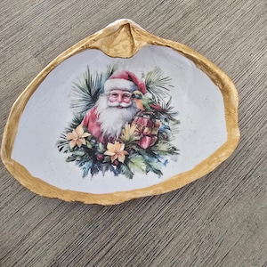 May include: A decorative seashell dish with a gold-painted rim. The interior features a festive image of Santa Claus, a parrot, and wrapped gifts surrounded by greenery and flowers. A unique holiday accent piece.