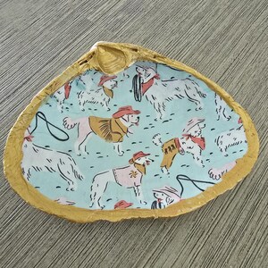 May include: A decorative seashell trinket dish with a gold-painted rim. The interior features a light blue background with a repeating pattern of cartoon dogs wearing cowboy hats and vests. A unique and whimsical home decor item.