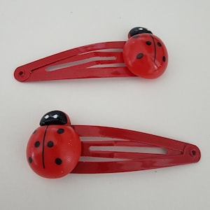 May include: Two red ladybug hair clips with black spots. Each clip has a metal bar with three teeth.