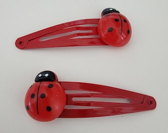 May include: Two red ladybug hair clips with black spots. Each clip has a metal bar with three teeth.