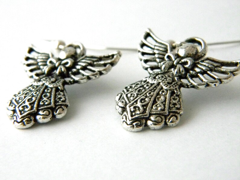 May include: A pair of silver angel earrings with intricate details. The angels have wings and a detailed dress with a bow at the neck.