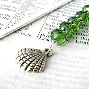 May include: A green glass bead bookmark with a silver seashell charm. The bookmark is on a page of a book with text.