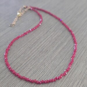 May include: A delicate, ruby-red beaded necklace with a gold-colored clasp and extender chain. The necklace is composed of small, faceted beads, creating a sparkling effect. The jewelry is displayed on a light-colored wooden surface.