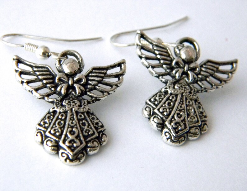 May include: A pair of silver angel dangle earrings. The angels have detailed wings and a bow at the neck. The earrings are on a white background.