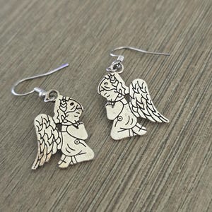 May include: A pair of silver-colored angel earrings. Each earring features a detailed angel design with wings, a halo, and a praying pose. The earrings have a hook closure and are displayed on a wooden surface.