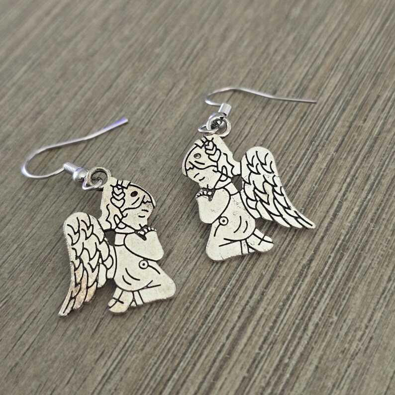 May include: A pair of silver-colored angel earrings. Each earring features a detailed angel design with wings, a halo, and a praying pose. The earrings have a hook closure and are displayed on a wooden surface.