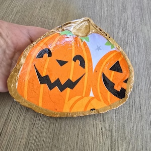 May include: A decorative ceramic dish shaped like a seashell with a gold rim. The dish is decorated with two orange pumpkins with black jack-o'-lantern faces.