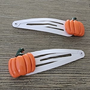 May include: Two white hair clips with orange pumpkin accents. The pumpkins have a green stem and are slightly raised from the clip.