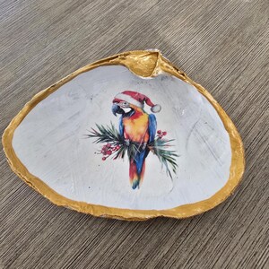 May include: A decorative seashell dish with a gold-painted rim. The interior is painted white and features a colorful parrot wearing a Santa hat, perched on a branch with red berries. A unique holiday-themed trinket dish.