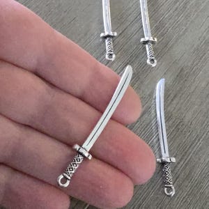 May include: Small silver-colored sword charms with detailed handles and curved blades. The charms have a loop at the base of the handle. The image shows several of these charms, suggesting they are for jewelry making or crafting.