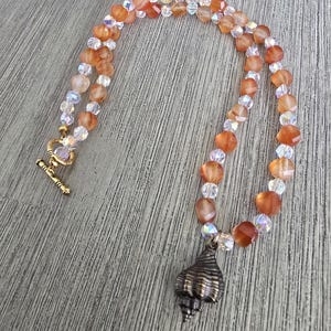 May include: A beaded necklace featuring alternating faceted orange carnelian beads and clear iridescent crystal beads. The necklace includes a gold-toned metal toggle clasp and a single bronze-toned metal seashell pendant hanging from the center.