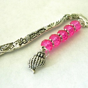 May include: Silver bookmark with a detailed design of a fish and floral accents. The bookmark has a pink crystal bead charm and a silver seashell charm.