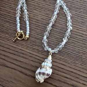 May include: A necklace featuring a series of clear, faceted, round beads strung together. The necklace includes a gold-toned toggle clasp and a pendant shaped like a seashell with white, tan, and brown stripes, accented with gold-toned metal details.