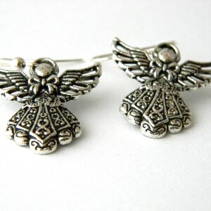 May include: A pair of silver angel earrings with intricate details. The angels have wings and a bow on their chest. The earrings are on a white background.