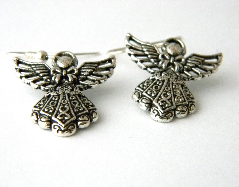 May include: A pair of silver angel earrings with intricate details. The angels have wings and a bow on their chest. The earrings are on a white background.