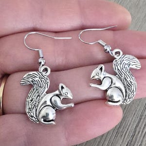 May include: Silver dangle earrings featuring a squirrel design. The squirrel is sitting with its tail curled up behind it.