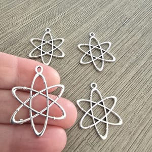 May include: Four silver-colored atom charm pendants. Each charm features a central nucleus with orbiting electrons, and a small loop for attaching to jewelry. The charms are displayed on a wooden surface.