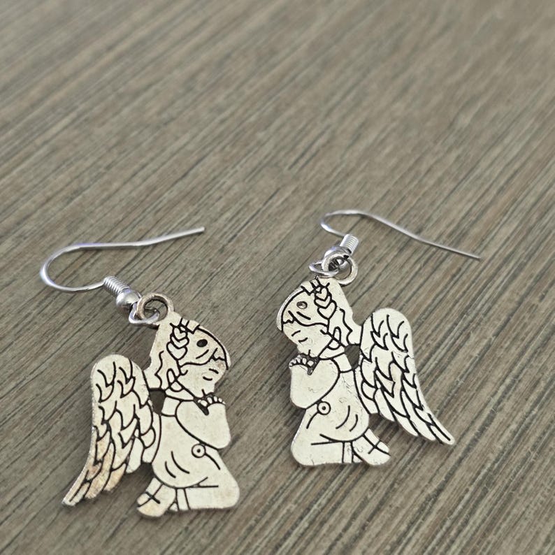 May include: A pair of silver-colored angel earrings. Each earring features a detailed angel design with wings, a halo, and clasped hands. The earrings have a hook closure.