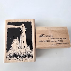 May include: Two rectangular wooden rubber stamps. One stamp features a black and white illustration of a lighthouse on a rocky shore. The other stamp displays the text: Thy WORD is a LAMP unto my FEET, and a LIGHT unto my PATH. -Psalms 119:105.