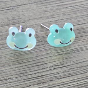 May include: A pair of light blue frog-shaped stud earrings. Each earring features a smiling frog face with black eyes and a light yellow patch on the cheeks. The earrings have silver posts.