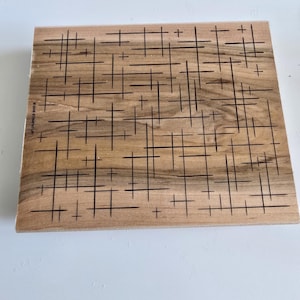 May include: A wooden block with a pattern of black lines and plus signs. The block is made of light brown wood and has a smooth surface. The text "©2002 STAMPIN' UP!" is printed on the block.