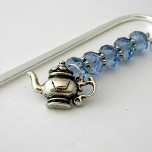 May include: A silver-toned bookmark featuring a small teapot charm and a strand of light blue faceted beads. The bookmark has a curved top and a decorative design, perfect for book lovers.