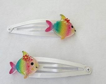 May include: Two white metal hair barrettes, each adorned with a colorful fish. The fish are rainbow-colored with pink tails and small red flower accents. The barrettes are on a white background.