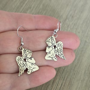 May include: A pair of silver-colored angel earrings. Each earring features a detailed angel figure with wings, a halo, and clasped hands. The earrings have a simple hook design for pierced ears.