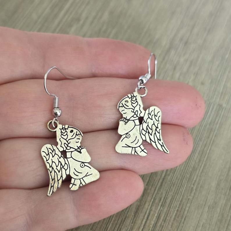 May include: A pair of silver-colored angel earrings. Each earring features a detailed angel figure with wings, a halo, and clasped hands. The earrings have a simple hook design for pierced ears.