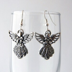 May include: Silver angel dangle earrings with intricate details and a bow on the chest. The earrings are hanging from a glass surface.